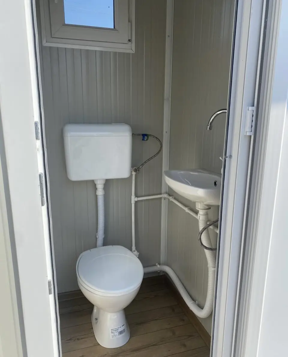 Inside a white flushing toilet unit with a ceramic-style toilet, a small sink and a frosted window