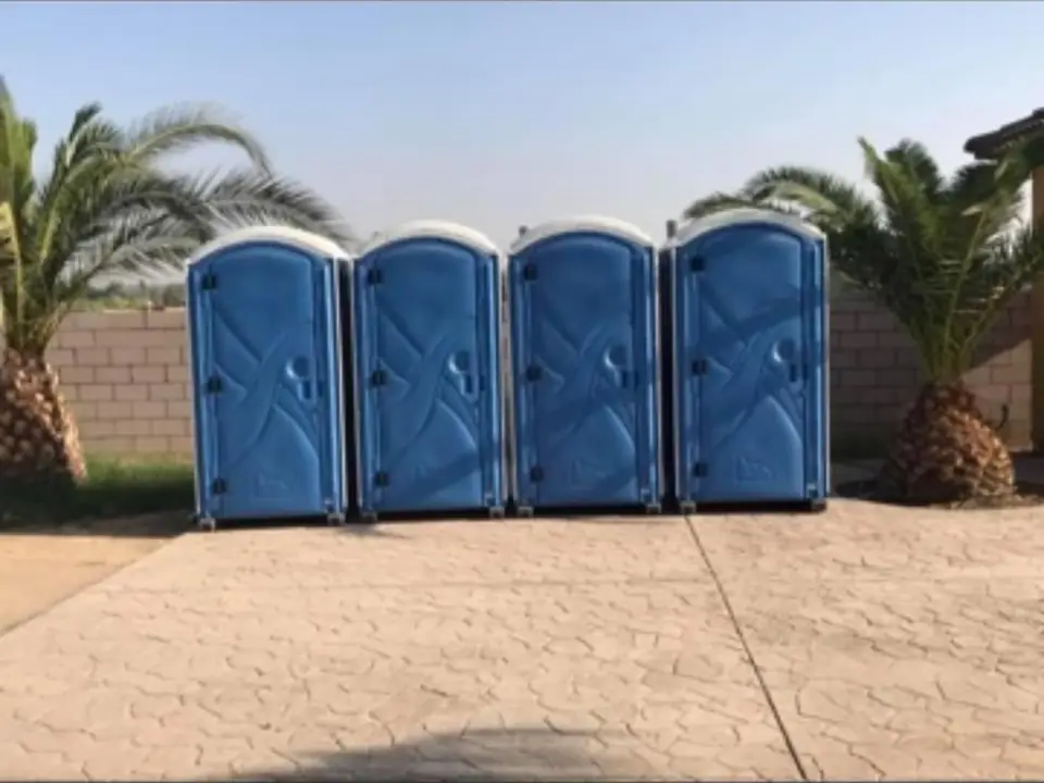 Four blue portable toilets lined up on a paved lot with palm trees behind them